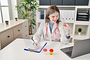 Young blonde woman doctor writing medical report holding urine test tube at clinic