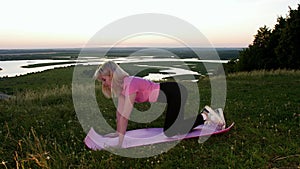 A young blonde woman doing exercises on yoga mat on sunset field - stretching her leg