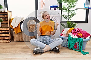 Young blonde woman doing laundry sitting by washing machine very happy and excited doing winner gesture with arms raised, smiling