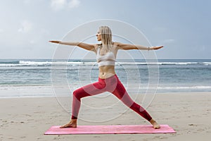Young blonde woman doing yoga in warrior pose in the beach. Pretty Female wearing sport clothes with sea at the background