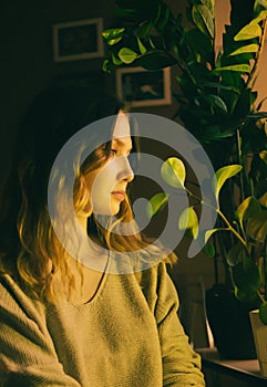 An young blonde woman dressed in comfortable home clothes sitting amidst houseplants in the evening light of a lamp. A cozy living