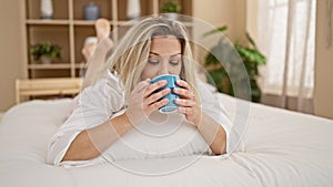 Young blonde woman drinking cup of coffee lying on bed at bedroom