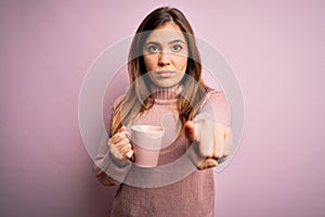 Young blonde woman drinking a cup of coffee over pink isolated background pointing with finger to the camera and to you, hand