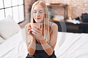 Young blonde woman drinking cup of coffee sitting on bed at bedroom