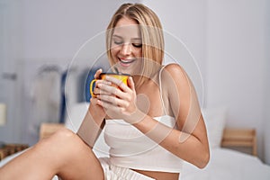 Young blonde woman drinking cup of coffee sitting on bed at bedroom