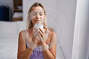 Young blonde woman drinking cup of coffee sitting on bed at bedroom