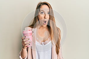 Young blonde woman drinking a take away cup of coffee scared and amazed with open mouth for surprise, disbelief face