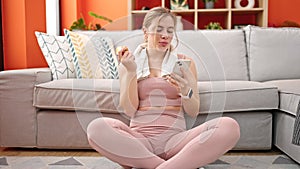 Young blonde woman eating apple using smartphone at home