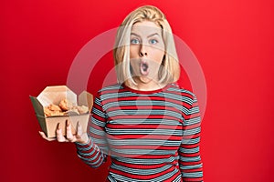 Young blonde woman eating fried chicken scared and amazed with open mouth for surprise, disbelief face