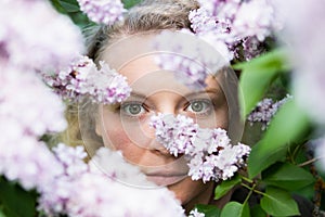 Young blonde woman face behind lilac bush in blossom