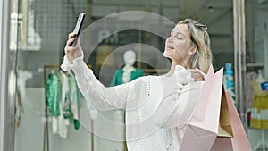 Young blonde woman going shopping holding bags making selfie by smartphone at street