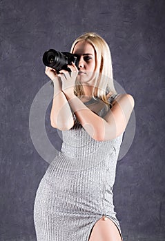 Young blonde woman in grey dress holding camera. Photographer making pictures.