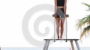 A young blonde woman gymnast uses high bars on the beach for her training - leaning on the stands and raising her feet