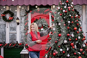 Young blonde woman hangs red balls on Christmas tree on street. Happy girl laughing in backyard