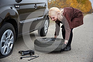 A young blonde woman hold spare tire near her car with a flat tire