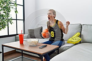 Young blonde woman holding basketball ball cheering tv game annoyed and frustrated shouting with anger, yelling crazy with anger