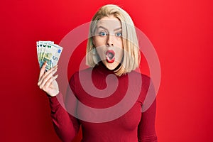 Young blonde woman holding bunch of 5 euro banknotes scared and amazed with open mouth for surprise, disbelief face