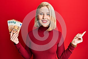 Young blonde woman holding bunch of 50 euro banknotes smiling happy pointing with hand and finger to the side