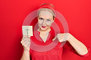 Young blonde woman holding catholic cross reminder pointing finger to one self smiling happy and proud