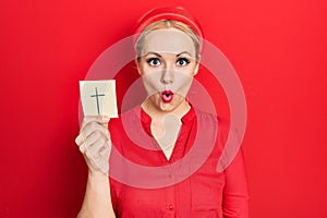 Young blonde woman holding catholic cross reminder scared and amazed with open mouth for surprise, disbelief face