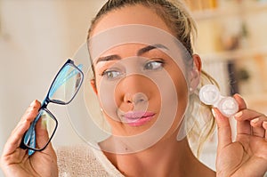 Young blonde woman holding contact lens case on hand and holding in her other hand a blue glasses on blurred background