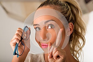 Young blonde woman holding contact lens on finger in front of her face and holding in her other hand a blue glasses on