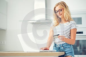Young blonde woman holding credit card and using laptop computer in home kitchen