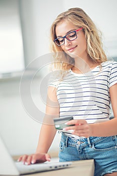 Young blonde woman holding credit card and using laptop computer in home kitchen