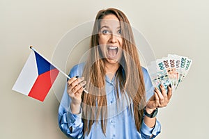Young blonde woman holding czech republic flag and koruna banknotes celebrating crazy and amazed for success with open eyes