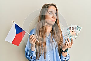 Young blonde woman holding czech republic flag and koruna banknotes smiling looking to the side and staring away thinking