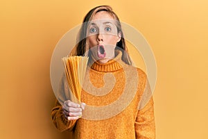 Young blonde woman holding dry spaghetti scared and amazed with open mouth for surprise, disbelief face