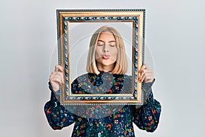 Young blonde woman holding empty frame looking at the camera blowing a kiss being lovely and sexy