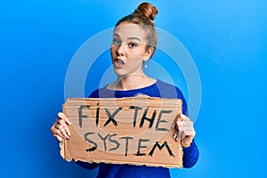 Young blonde woman holding fix the system banner cardboard in shock face, looking skeptical and sarcastic, surprised with open