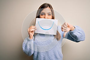 Young blonde woman holding funny smile drawing on mouth as happy expression with angry face, negative sign showing dislike with