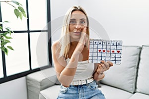 Young blonde woman holding heart calendar hand on mouth telling secret rumor, whispering malicious talk conversation