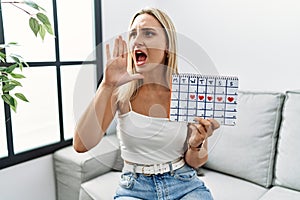 Young blonde woman holding heart calendar shouting and screaming loud to side with hand on mouth