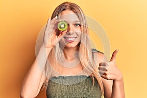 Young blonde woman holding kiwi smiling happy and positive, thumb up doing excellent and approval sign