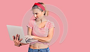 Young blonde woman holding laptop thinking attitude and sober expression looking self confident