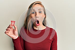 Young blonde woman holding menstrual cup scared and amazed with open mouth for surprise, disbelief face