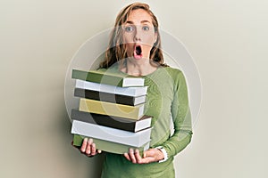 Young blonde woman holding a pile of books afraid and shocked with surprise and amazed expression, fear and excited face
