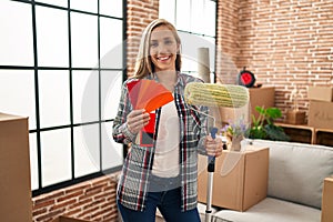 Young blonde woman holding roller painter and paint samples at new home smiling with a happy and cool smile on face