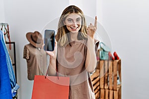 Young blonde woman holding shopping bags showing smartphone screen smiling with an idea or question pointing finger with happy