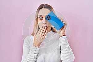 Young blonde woman holding smartphone covering eye covering mouth with hand, shocked and afraid for mistake