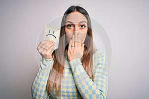 Young blonde woman holding sticky paper note with sad face emoticon over isolated background cover mouth with hand shocked with