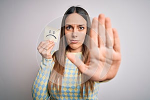 Young blonde woman holding sticky paper note with sad face emoticon over isolated background with open hand doing stop sign with