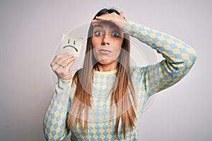 Young blonde woman holding sticky paper note with sad face emoticon over isolated background stressed with hand on head, shocked