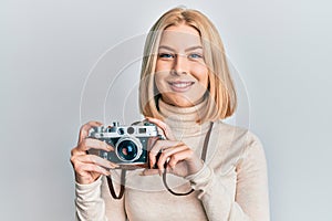Young blonde woman holding vintage camera looking positive and happy standing and smiling with a confident smile showing teeth