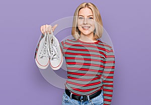 Young blonde woman holding white casual shoes looking positive and happy standing and smiling with a confident smile showing teeth