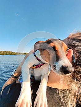 A young blonde woman holds a Jack Russell terrier dog on her shoulder. They are sailing in a boat on a wide river. Tourism and