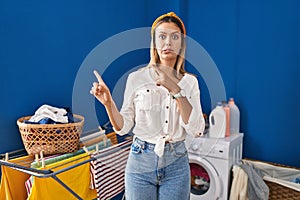 Young blonde woman at laundry room pointing aside worried and nervous with both hands, concerned and surprised expression